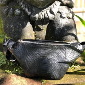 May include: A black fanny pack featuring a textured snakeskin pattern across the exterior. The bag has a curved silhouette with a silver-toned metal zipper closure running along the top edge and an adjustable strap attached to the sides.