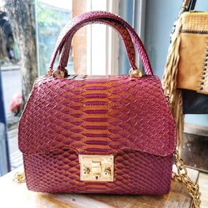 May include: A structured handbag featuring a textured, scale-like pattern in a vibrant magenta and copper gradient. The bag includes a single top handle, a gold-toned metal push-lock closure on the front flap, and a gold-toned chain shoulder strap.