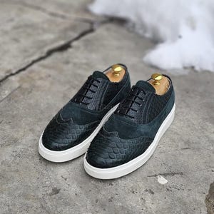 Puede incluir: Un par de zapatos negros con cordones que combinan cuero texturizado con patrón de reptil y paneles de ante. Los zapatos tienen un diseño de puntera tipo wingtip, cordones negros y una suela de goma blanca gruesa con horma de madera.
