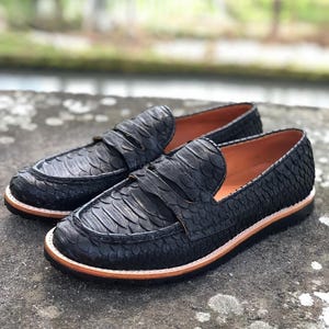 Könnte beinhalten: Ein Paar schwarze Slipper mit einem strukturierten, schlangenlederartigen Muster auf dem Obermaterial. Die Schuhe haben ein klassisches Slip-on-Design mit einem Riemen über dem Vorderblatt, einem hellbraunen Innenfutter und einer dicken schwarzen Sohle.