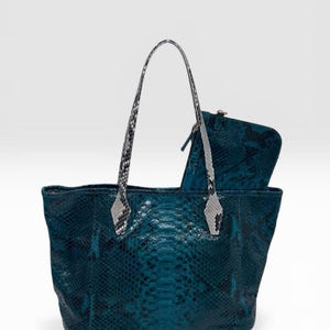 May include: A teal tote bag with a dark snakeskin pattern and two tall, light gray and black snakeskin-textured handles. A matching rectangular zippered pouch is tucked inside the bag, featuring the same teal and dark snakeskin texture.
