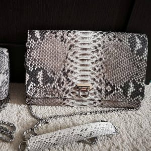 May include: A rectangular clutch bag featuring a grey, white, and black snakeskin pattern. The bag has a silver-toned metal clasp closure on the front flap and a detachable silver chain shoulder strap with a matching snakeskin leather shoulder pad.