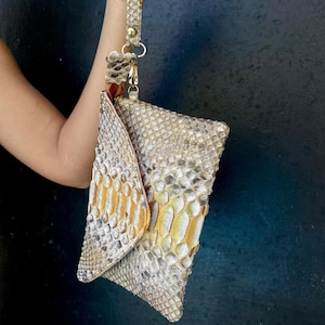 Peut inclure: Une pochette avec dragonne pr&eacute;sentant un motif textur&eacute; en peau de serpent dans des tons beige, gris et or m&eacute;tallique. Le sac poss&egrave;de un rabat triangulaire et une dragonne assortie fix&eacute;e par un fermoir et des &eacute;l&eacute;ments en m&eacute;tal dor&eacute;.