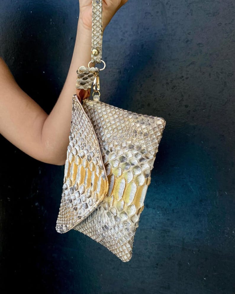Peut inclure: Une pochette avec dragonne pr&eacute;sentant un motif textur&eacute; en peau de serpent dans des tons beige, gris et or m&eacute;tallique. Le sac poss&egrave;de un rabat triangulaire et une dragonne assortie fix&eacute;e par un fermoir et des &eacute;l&eacute;ments en m&eacute;tal dor&eacute;.