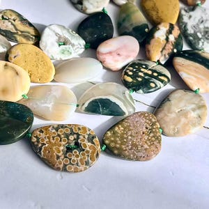 May include: A collection of irregularly shaped, polished stone beads in various colors including mustard yellow, forest green, cream, and soft pink. The stones feature natural patterns, including spotted and banded textures, and are strung on a thin green cord.