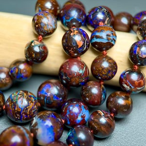 May include: A strand of round, polished boulder opal beads featuring a dark brown matrix with vibrant, iridescent flashes of blue, green, and orange. The beads are strung together on a thin red cord, showcasing natural color variations.