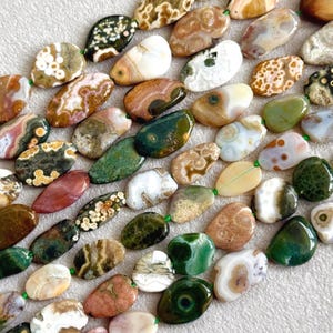 Puede incluir: Una colección de cuentas de piedras preciosas pulidas, de forma irregular y de lágrima, en varios tonos tierra que incluyen verde musgo, crema, beige, marrón y blanco. Las piedras presentan patrones naturales como manchas, remolinos y bandas.