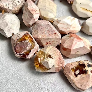 May include: A collection of faceted, irregular beads featuring a mix of pale pink, cream, and white stone textures. Several beads display translucent, amber-colored crystalline inclusions within their centers, strung together on a thin cord.