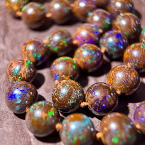 May include: A necklace composed of round, polished boulder opal beads. Each bead features a dark brown base color with vibrant, iridescent flashes of green, blue, and purple light reflecting from within the stone. The beads are strung together.