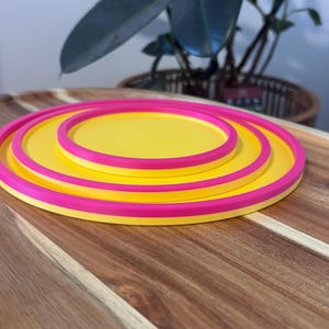 May include: A set of three nested circular coasters featuring a bright yellow center and a vibrant hot pink border. The coasters are stacked in descending order of size, creating a layered appearance with consistent color contrast throughout.