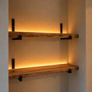 May include: Two horizontal shelves made of textured, natural-toned wood, each supported by black metal brackets. The shelves feature integrated warm LED lighting strips along the back edge, casting a soft glow against the wall behind them.