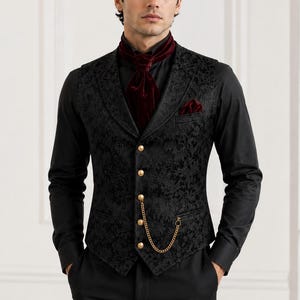 Könnte beinhalten: Eine schwarze Weste mit tonalen Blumenmustern, fünf goldenen Knöpfen und einer goldenen Taschenuhrkette. Die Weste wird über einem schwarzen Langarmhemd mit einem dunkelroten Samt-Plastron und einem passenden Einstecktuch getragen.