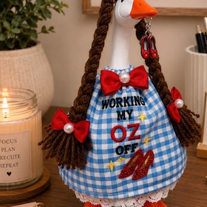 May include: A white goose figurine with brown yarn braids, wearing a blue and white gingham dress with a red bow and white lace trim. The dress features the text "WORKING MY OZ OFF" and a graphic of red glittery shoes. A pair of red shoe charms hangs from the beak.