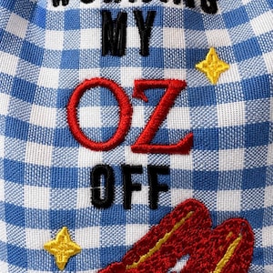 May include: A blue and white gingham fabric pouch featuring black embroidered text that reads WORKING MY OFF with the red embroidered word OZ in the center. The design includes two yellow embroidered stars and a pair of red slippers.