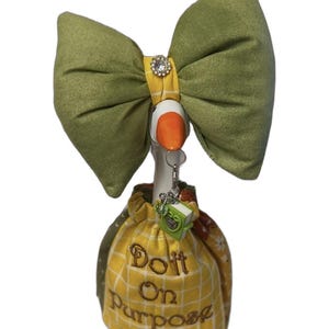 May include: A decorative goose figurine wearing a large olive green fabric bow with a central rhinestone accent. The goose is dressed in a yellow and white grid-patterned fabric outfit featuring the embroidered text "Do It On Purpose".