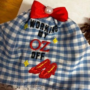 May include: A blue and white gingham fabric item featuring a red ribbon bow with a white pearl-like bead at the top. The text "WORKING MY OZ OFF" is embroidered in black and red, accompanied by yellow star icons and red shoe embroidery.