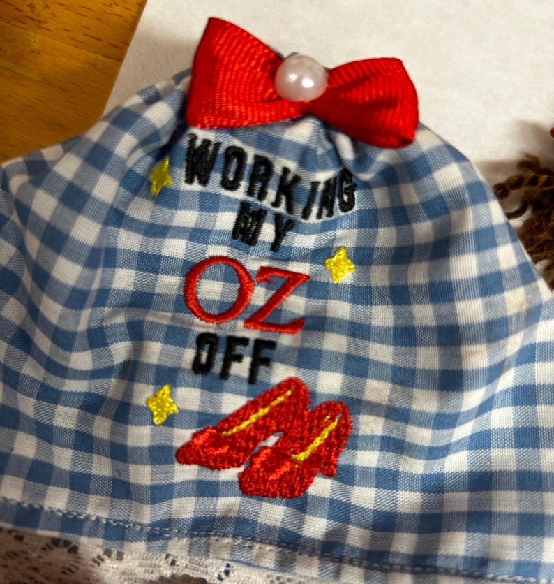 May include: A blue and white gingham fabric item featuring a red ribbon bow with a white pearl-like bead at the top. The text "WORKING MY OZ OFF" is embroidered in black and red, accompanied by yellow star icons and red shoe embroidery.