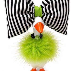 May include: A white goose figurine featuring an orange beak and feet, wearing a large black and white striped fabric bow with a lime green center, a lime green feather boa collar, and a necklace with black beads and a lime green rhinestone sphere.