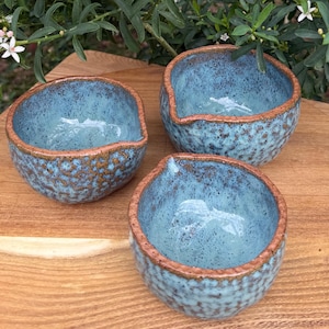 May include: Three small ceramic bowls with a light blue speckled glaze and a textured, reddish-brown rim. Each bowl features a distinct pouring spout on one side. The exterior surfaces have a dimpled, tactile pattern, and the interior is smooth.