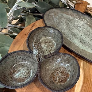 May include: A three-section ceramic serving dish with a central handle and a separate elongated oval tray. Both pieces feature a speckled light green and grey glaze with dark brown, textured, uneven edges and a rustic, hand-formed appearance.