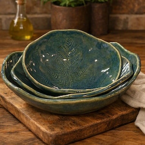 May include: A set of three nested ceramic bowls with an organic, wavy rim design. The bowls feature a deep teal and green glaze with a prominent, detailed fern leaf impression embossed in the centre of each piece, creating a textured surface.