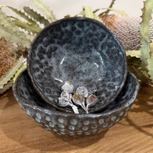 May include: Two nested ceramic bowls with a dark, mottled blue and black glaze. The exterior of the larger bowl features a textured, dimpled surface, while the interior of both bowls shows a smooth, glossy finish with dark speckles throughout.