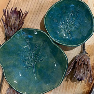May include: Two irregular, teal-coloured ceramic dishes with glossy finishes, each featuring a detailed botanical leaf imprint in the centre. The dishes are accompanied by two dried, brownish-purple protea flowers with pointed, layered petals.