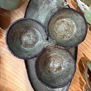 May include: A set of three small, round ceramic bowls attached to an elongated, oval-shaped ceramic tray. The pottery features a speckled, light green and grey glaze with dark brown, textured, uneven rims and a glossy finish throughout.