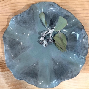 May include: A round, hand-formed ceramic bowl with a wavy, scalloped edge and a glossy, mottled blue-grey glaze. A small, dried eucalyptus sprig with pale green leaves and dark seed pods rests in the centre of the bowl's smooth, concave surface.
