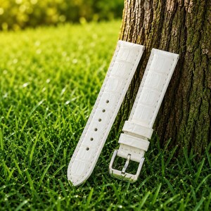 Pode incluir: Uma bracelete de relógio branca com um padrão texturizado de imitação de crocodilo. A bracelete é composta por duas peças, uma com uma série de orifícios de ajuste e a outra com uma fivela de metal prateado e dois passadores de pele branca.