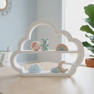 May include: A white cloud-shaped shelf with two tiers. The top shelf holds a pink and white yarn ball, a small light green bear figurine, and a pastel rainbow. The bottom shelf features a small blue cloud figurine and a crescent moon shape.