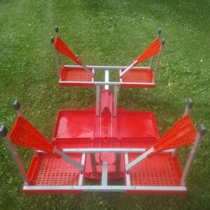 May include: A red folding table with four chairs. The table and chairs are made of metal and plastic. The table is folded up and the chairs are attached to the table.