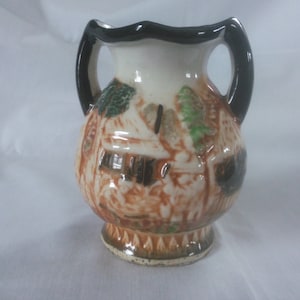 May include: A small, white ceramic vase with black handles and a brown and green painted design of a village scene.