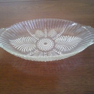 May include: A clear glass serving dish with a scalloped edge and a decorative etched design of leaves and circles. The dish has two handles on the sides.