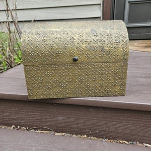 May include: A small, antique-style chest with a domed lid. The chest is a muted gold color and covered in a repeating floral pattern. A small, dark knob serves as the latch. The chest is sitting on a brown surface.