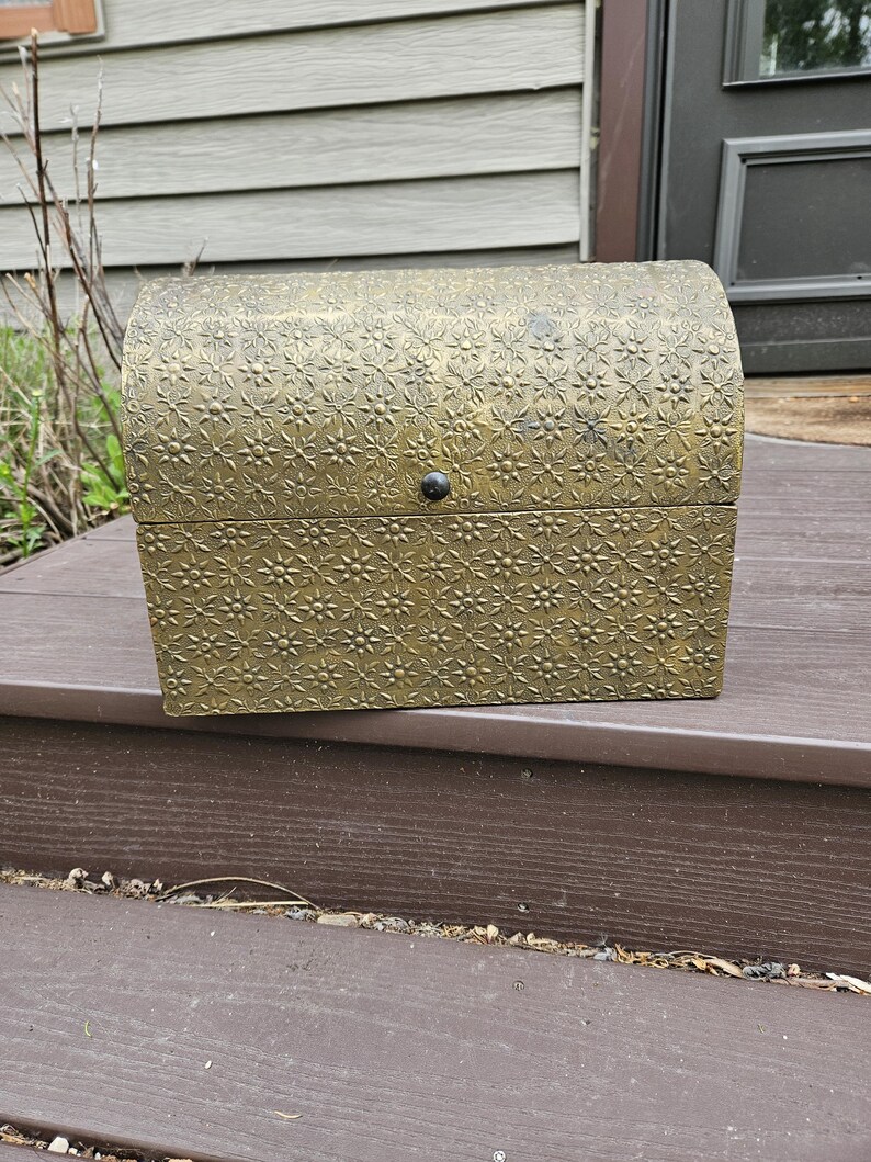 May include: A small, antique-style chest with a domed lid. The chest is a muted gold color and covered in a repeating floral pattern. A small, dark knob serves as the latch. The chest is sitting on a brown surface.