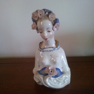 May include: A porcelain figurine of a woman with a serene expression. The bust features a white dress with pink floral accents and blue trim. Her hair is adorned with pink flower accents and a blue headpiece. A pink rose is held in her hands.