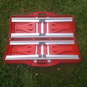 May include: Red folding table and chairs in a closed position. The table and chairs are made of red plastic with silver metal supports. The table and chairs are folded up and ready to be transported.