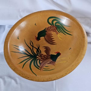 May include: A wooden bowl with a hand-painted design of two roosters facing each other. The roosters are painted in shades of brown, green, and black, with yellow accents. The bowl is a light brown color and has a smooth, rounded surface.