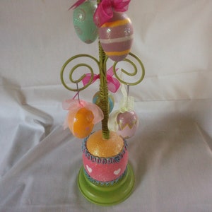 May include: A green metal Easter egg tree stand with a pink and orange glitter base. The stand is decorated with pastel colored Easter eggs and pink ribbons.