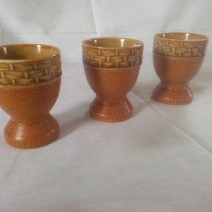 May include: Three brown ceramic egg cups with a woven pattern around the top.