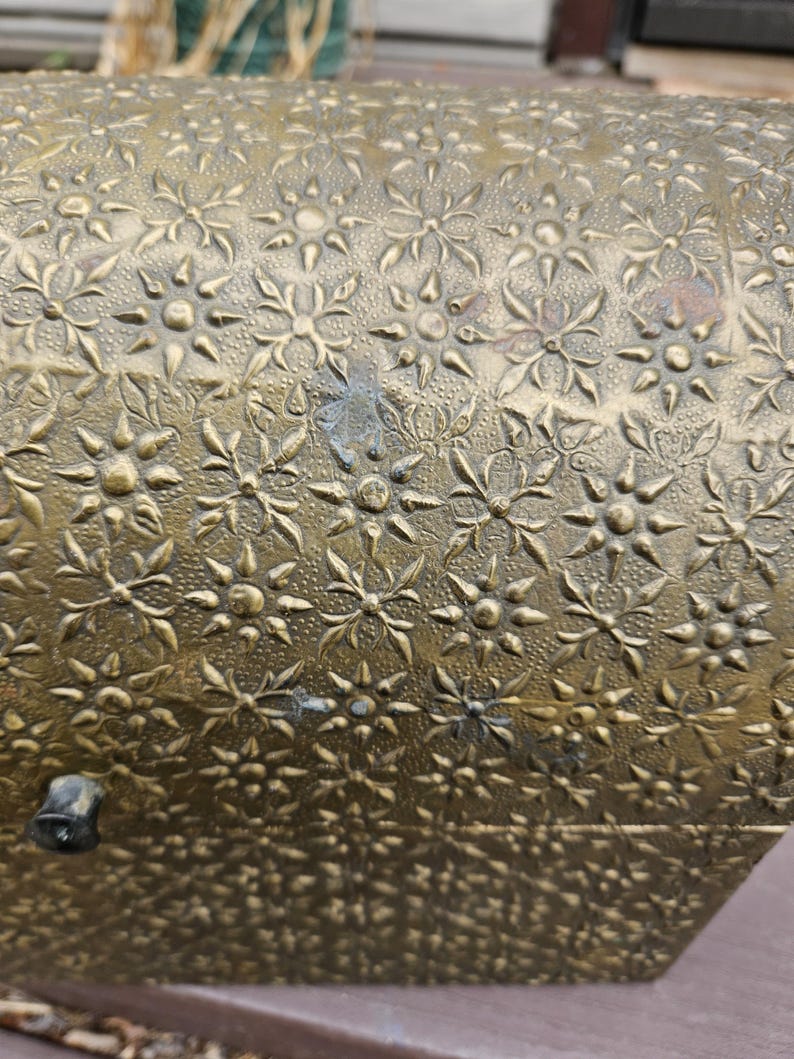 May include: Close-up of a vintage brass box with a detailed floral pattern. The box has a textured surface with embossed flowers and leaves. The brass has a warm, aged patina, and a small latch is visible on the side. The box is likely used for storage.