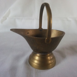May include: A small, antique brass-colored metal container with a handle. The container has a spout and a rounded base with a decorative pattern. The handle is arched, and the overall design suggests a vintage or decorative item.