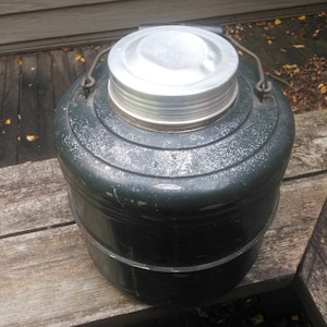 May include: A vintage green metal thermos with a silver lid. The thermos has a handle and a textured surface.