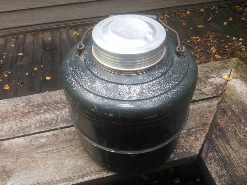 May include: A vintage green metal thermos with a silver lid. The thermos has a handle and a textured surface.