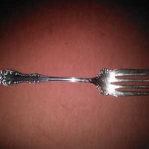 May include: A silver fork with an ornate handle and three tines. The fork is on a brown surface.