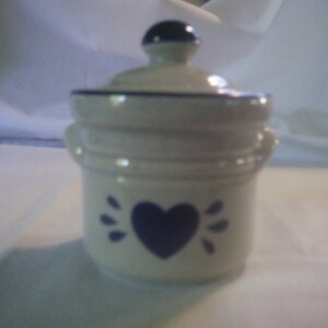May include: A small white ceramic jar with a blue lid and a blue heart design. The jar has a blue rim and a blue band around the middle.