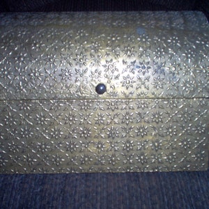 May include: A small, antique-style chest with a domed lid. The chest is made of a metallic material with a floral pattern embossed across its surface. A small, dark knob serves as the latch.