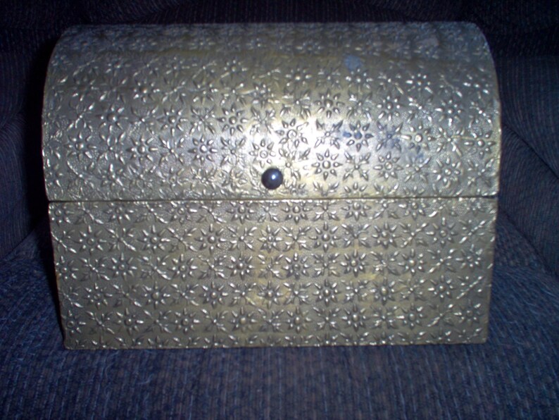 May include: A small, antique-style chest with a domed lid. The chest is made of a metallic material with a floral pattern embossed across its surface. A small, dark knob serves as the latch.