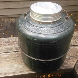 May include: A vintage green metal water cooler with a silver lid. The cooler has a rounded shape and a slightly textured surface.